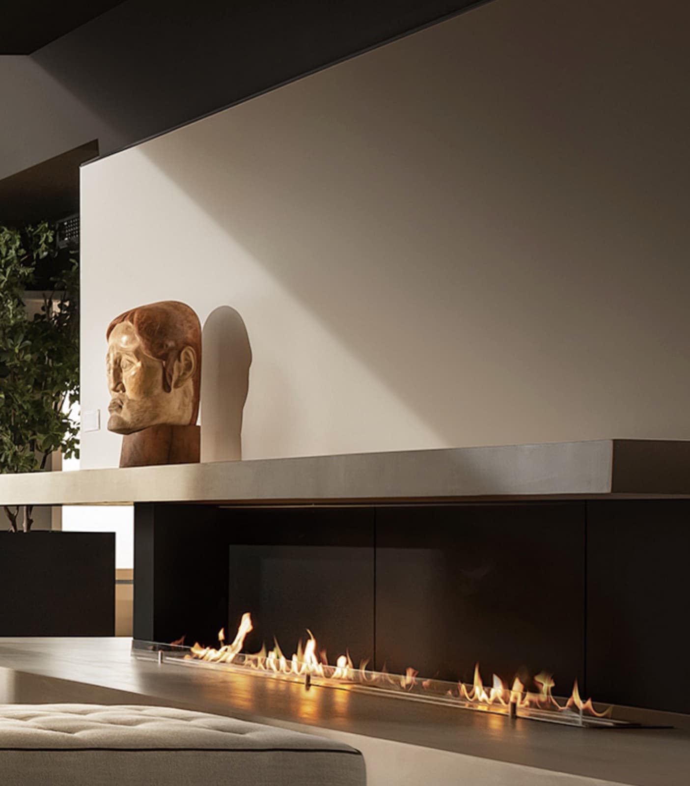 A linear fireplace set into a dark hearth beneath a concrete shelf, with a carved wooden head and a fig tree beside it